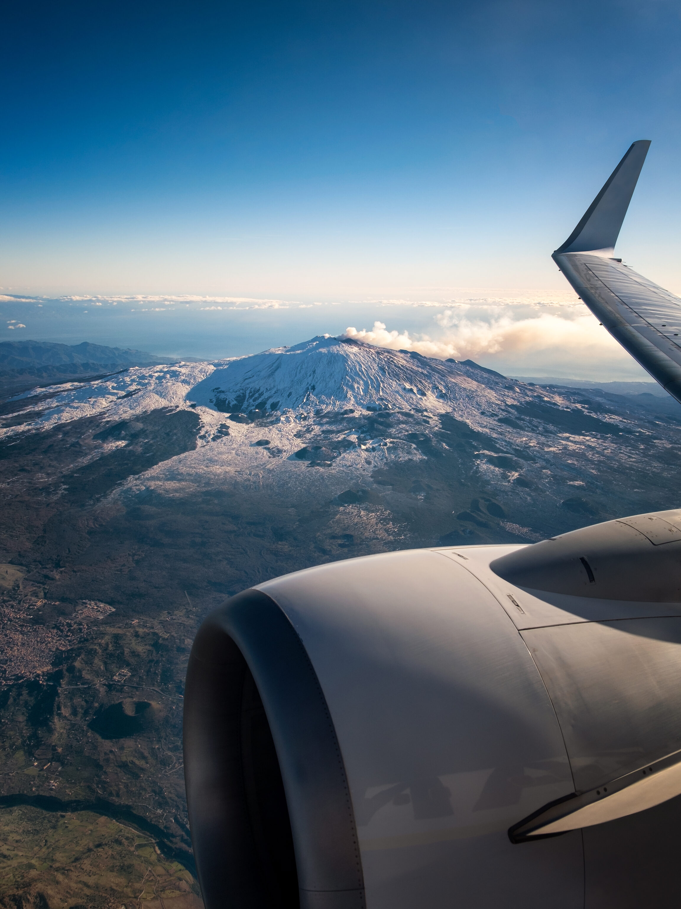Snow-covered Mount Etna seen from an aircraft window, with the wing and engine in the foreground